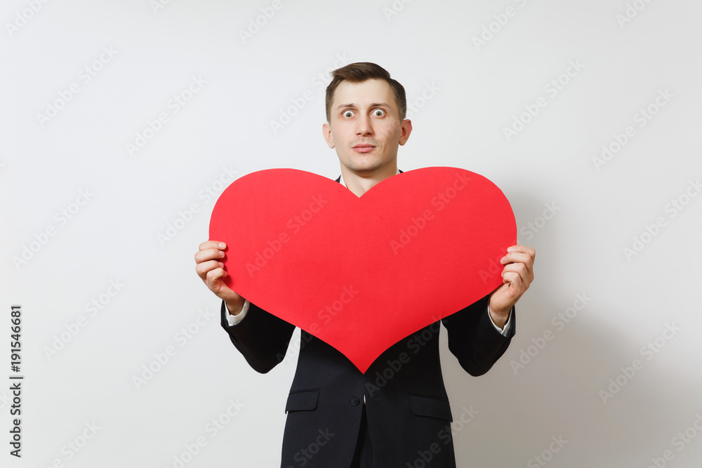Young stunned shocked upset sad man in suit holding big red heart isolated on white background. Copy space for advertisement. St. Valentine's Day, International Women's Day, birthday, holiday concept. Young stunned shocked upset sad man in suit holding big red heart isolated on white background. Copy space for advertisement. St. Valentine's Day, International Women's Day, birthday, holiday concept.