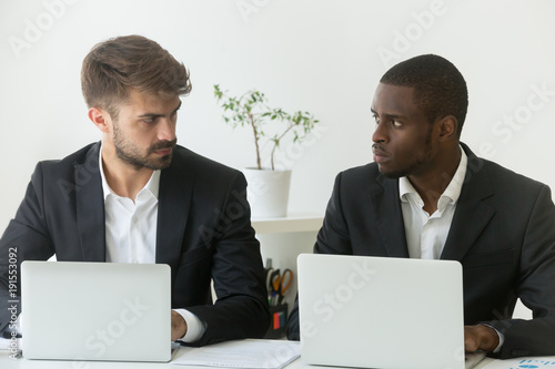 Fotografia Multiracial office rivals looking at each other with hate envy sitting with lapt