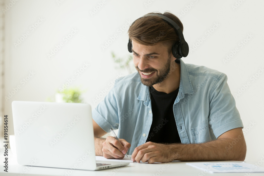 Smiling man in headphones looking at computer screen watching webinar, making business video call, young businessman consulting remote client online writing notes, hr holding distance job interview Smiling man in headphones looking at computer screen watching webinar, making business video call, young businessman consulting remote client online writing notes, hr holding distance job interview