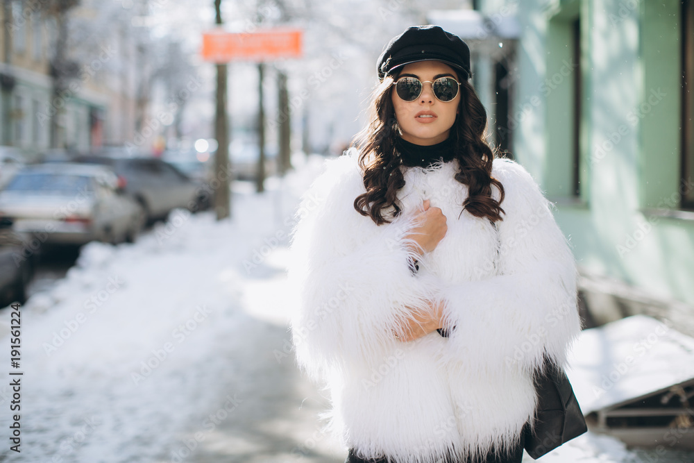A beautiful, stylish, fashionable woman in a fur coat, hat and glasses, posing on the street in snowy weather.