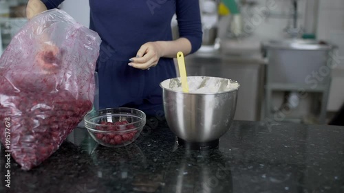 Woman from large package on kitchen table lays out frozen berries. For dish of cherries female chef prepares ingredients, which she puts from transparent bag into metal bowl after that washes hands in photo