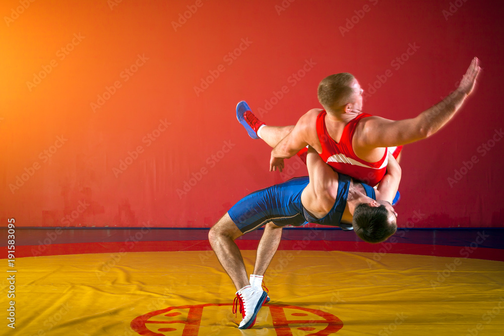 Two greco-roman wrestlers in red and blue uniform making a suplex wrestling on a yellow wrestling carpet in the gym Two greco-roman wrestlers in red and blue uniform making a suplex wrestling on a yellow wrestling carpet in the gym