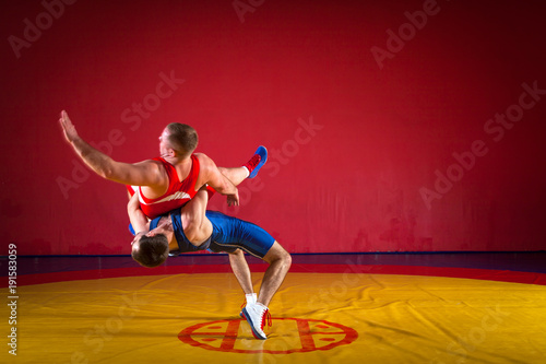 Two greco-roman wrestlers in red and blue uniform making a suplex wrestling on a yellow wrestling carpet in the gym