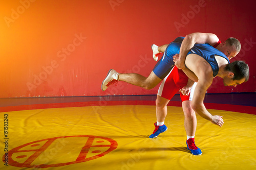 Two greco-roman wrestlers in red and blue uniform wrestling on background on a yellow wrestling carpet in the gym