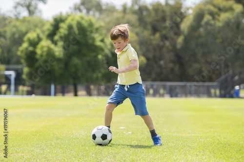 little kid 7 or 8 years old enjoying happy playing football soccer at grass city park field running and kicking the ball excited in childhood sport passion