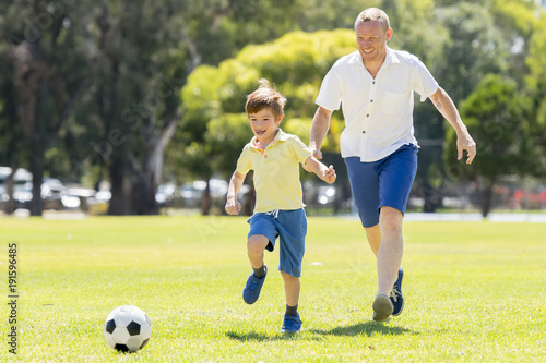 young happy father and excited little 7 or 8 years old son playing together soccer football on city park garden running on grass kicking the ball