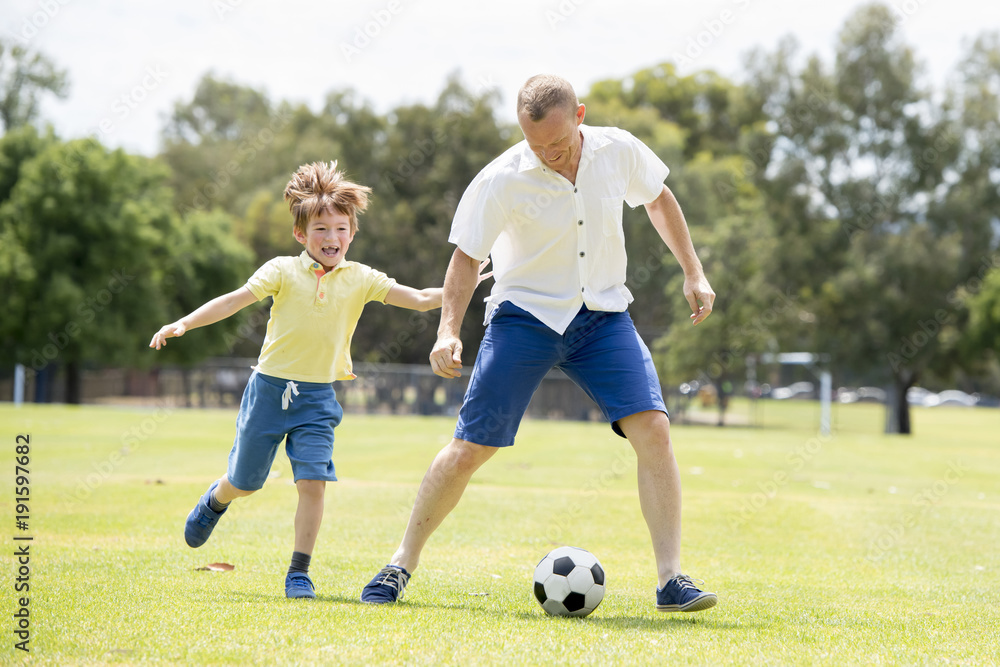 Obraz premium young happy father and excited little 7 or 8 years old son playing together soccer football on city park garden running on grass kicking the ball