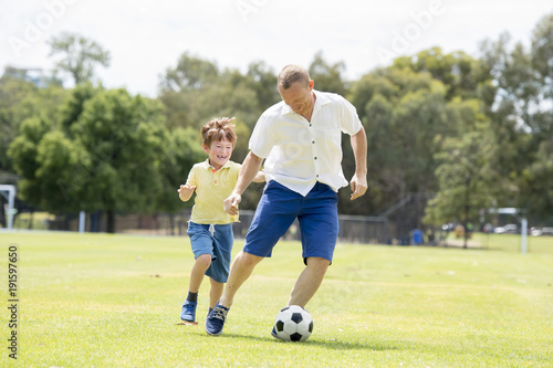 young happy father and excited little 7 or 8 years old son playing together soccer football on city park garden running on grass kicking the ball