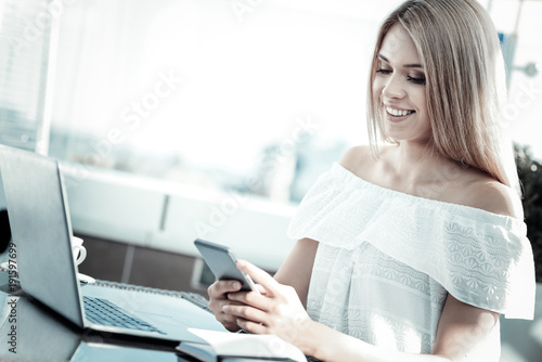 Always online. Delighted positive young woman sitting at the table and holding her smartphone while using social media