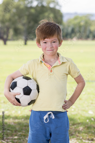 young little kid 7 or 8 years old enjoying happy playing football soccer at grass city park field posing smiling proud standing holding the ball