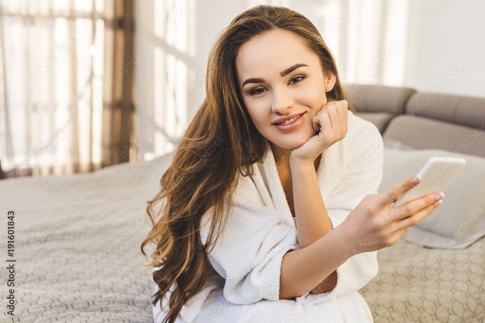 smiling young woman in bathrobe with smartphone sitting on bed smiling young woman in bathrobe with smartphone sitting on bed