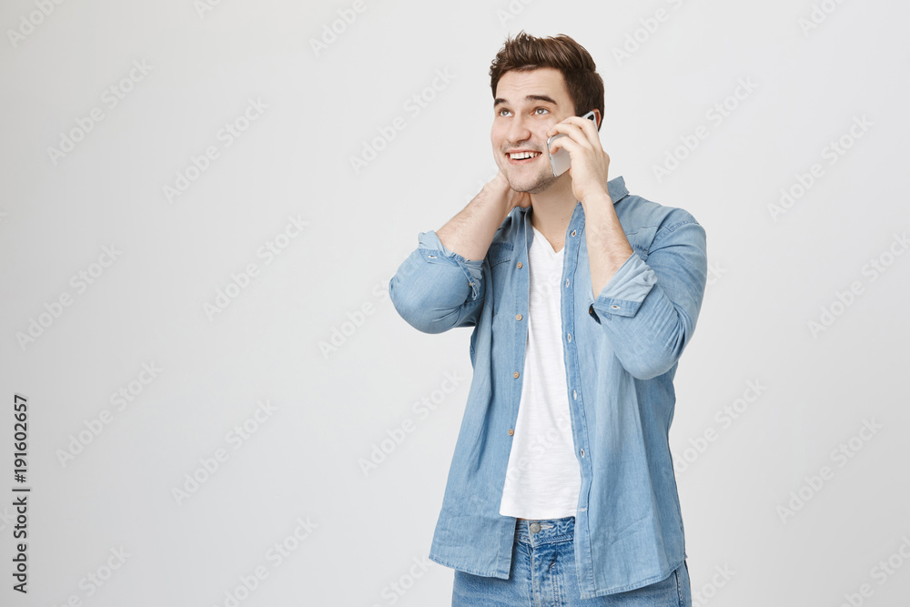 Portrait of young handsome man with stylish haircut, wearing denim shirt, speaking on smartphone while touching his neck and smiling broadly, isolated over white background. Portrait of young handsome man with stylish haircut, wearing denim shirt, speaking on smartphone while touching his neck and smiling broadly, isolated over white background.