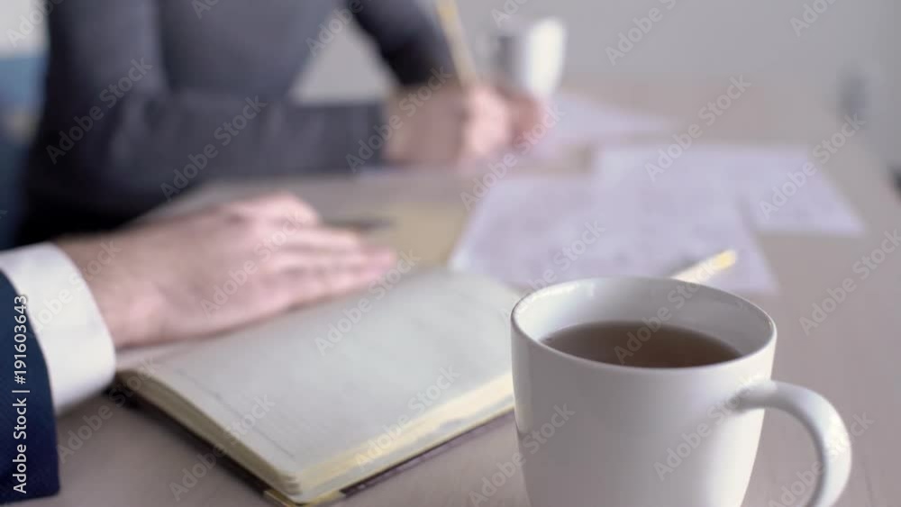 White cup with tea on background of working people at the desk. During work man's hand in suit takes a mug with a drink to quench his thirst with a continuous brainstorming session with co-workers at White cup with tea on background of working people at the desk. During work man's hand in suit takes a mug with a drink to quench his thirst with a continuous brainstorming session with co-workers at