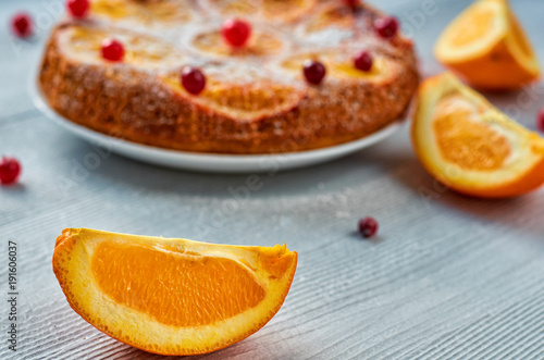 Juicy orange slice on the foreground close up with free copy space. Homemade orange cake with fresh red cranberries and oranges on the blurred background. Citrus cake on the gray table. Side view