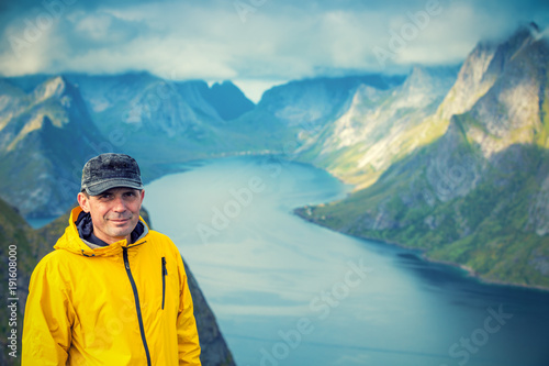Portrait of a tourist man, standing on a peak of a mount Reinebringen and looking in camera. Beautiful mountain landscape in the background. Nature of Norway, Lofoten Islands