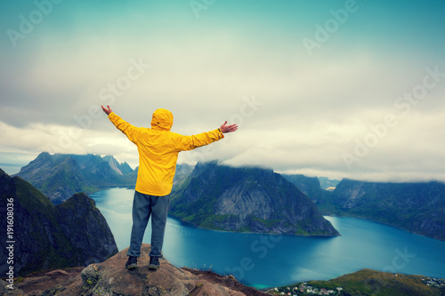 Panoramic aerial view of the fjord from the mountain. Man with hands in the air standing on a cliff of mount Reinebringen. Beautiful mountain landscape. Nature Norway, Lofoten islands, Reine 