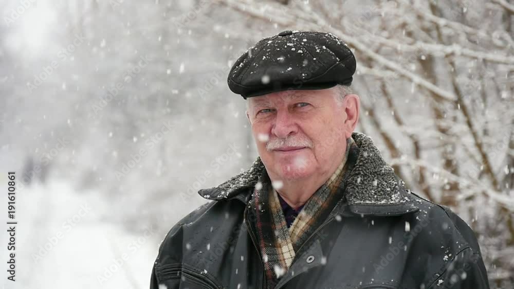 An amazing view of a senior man with white mustashe in a flat cap. He smiles and stands in a snowy park in winter. The snowflakes whirl over him. An amazing view of a senior man with white mustashe in a flat cap. He smiles and stands in a snowy park in winter. The snowflakes whirl over him.