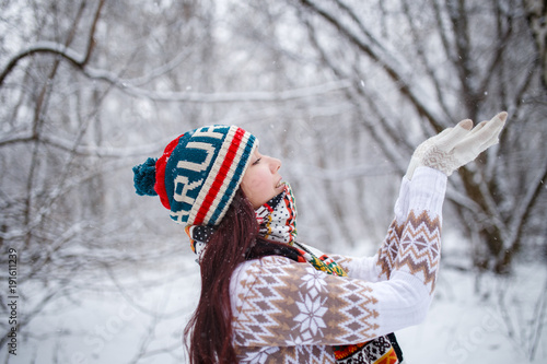 Photo of smiling girl in knitted hat and scarf catching snowflakes in winter forest during day