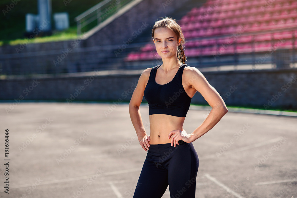 Beautiful athletic girl with fit body in black sportswear is resting after  training. Sporty girl with perfect body on the stadium doing workout. Stock  Photo