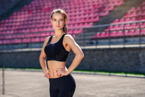 Beautiful athletic girl with fit body in black sportswear is resting after training. Sporty girl with perfect body on the stadium doing workout.