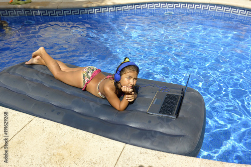 A girl in bikini with computer and headphones sunbathing on an inflatable mattress in the pool