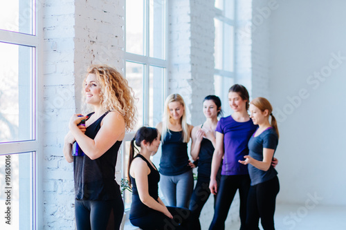Close up image of attractive curly blond fitness woman over blurred group of people in white gym interior. Teamwork, good mood and healthy lifestyle concept.