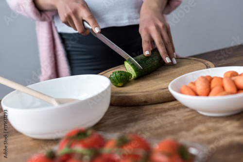 cropped view of woman cutting cucumber and making salad at kitchen
