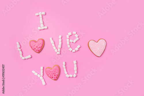 Marshmallows on pink background with sign in English I Love You. Flat lay or top view. Background or texture of colorful mini marshmallows. Gingerbread hearts. Valentines day background concept.