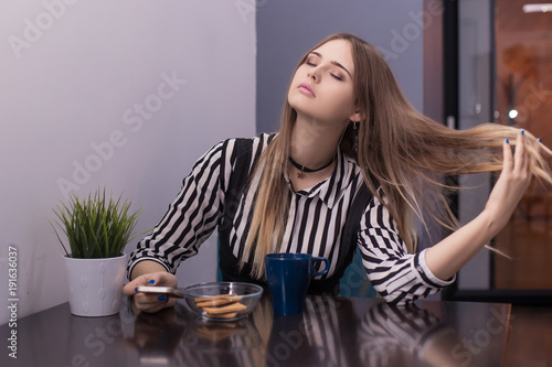 A beautiful young woman sits at a table with a mug of tea and a biscuit straightens, throws back her hair, holds the phone in her hand and smiles. Flirt, radiate the pheramons.