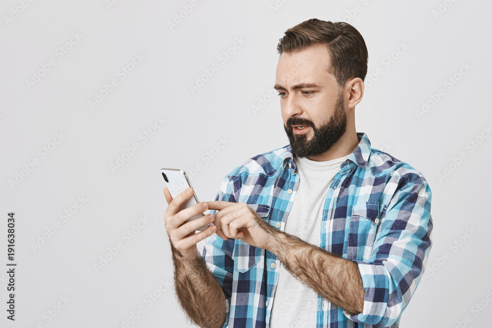 Portrait of attractive guy with beard and moustache holding smartphone and browsing network with surprised and perplexed expression, standing against gray background. Husband just got anxious message Portrait of attractive guy with beard and moustache holding smartphone and browsing network with surprised and perplexed expression, standing against gray background. Husband just got anxious message