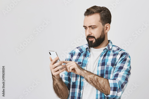 Portrait of attractive guy with beard and moustache holding smartphone and browsing network with surprised and perplexed expression, standing against gray background. Husband just got anxious message
