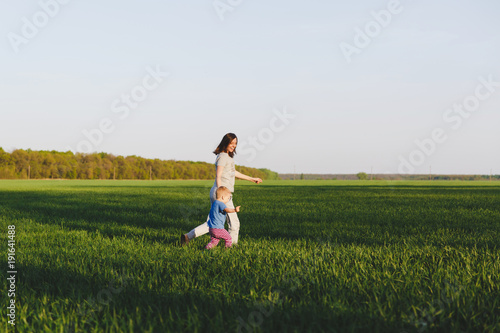 Joyful young woman walk on green grass field wheat background, rest, have fun, play, run with little cute child baby boy. Mother, small kid son. Family day 15 of may, love, parents, children concept.