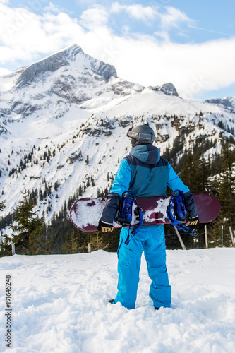 Junger Snowboarder steht am Rande der Piste und blickt auf die Berge