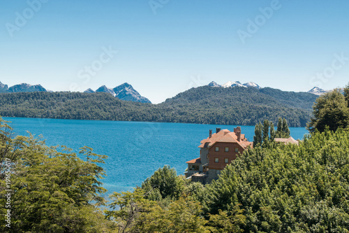 Brick house facing a blue lake, in wooded area and snowy mountains in the background