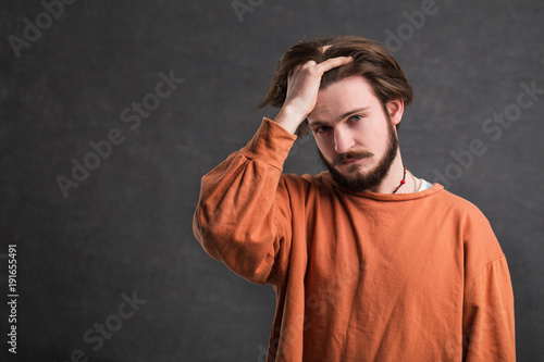 Portrait of serious, handsome man with a dark beard, wearing casual orange shirt, stressed and tired, indoor shot in the gray background