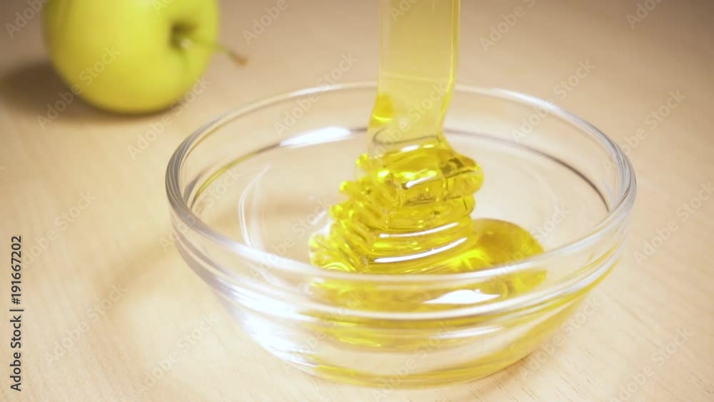Slow motion transparent honey flows into a glass bowl on the table next to the apple Slow motion transparent honey flows into a glass bowl on the table next to the apple