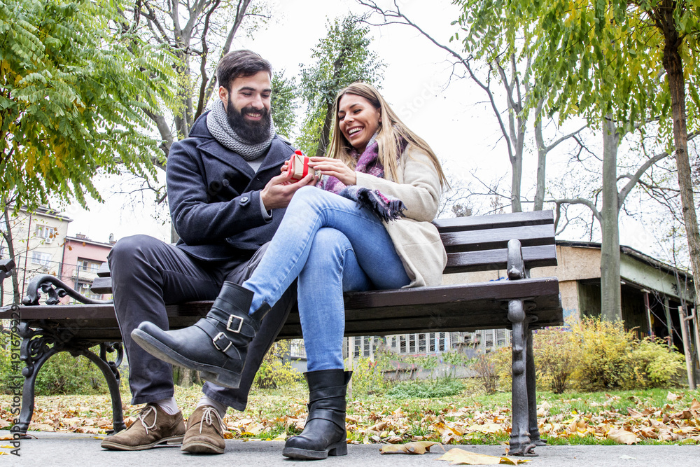 Beautiful young couple sitting on the bench in park and guy giving a small box to his girfriend as a gift and she smile Beautiful young couple sitting on the bench in park and guy giving a small box to his girfriend as a gift and she smile