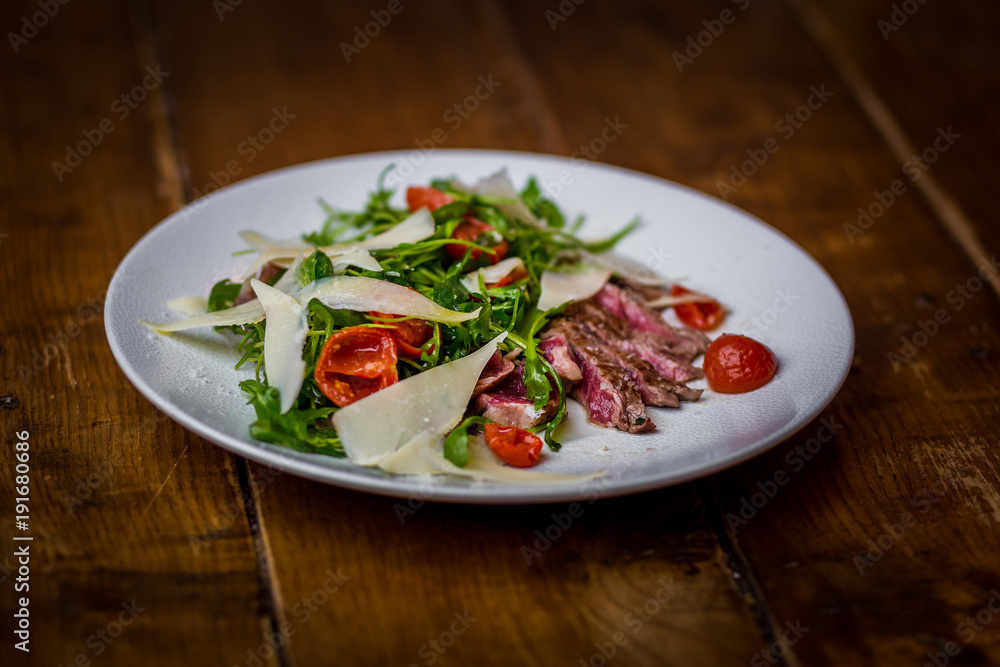 top view of italian sliced beef salad with rucola, tomatoes and parmesan cheese on white plate on wooden table