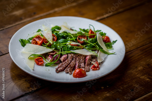 top view of italian sliced beef salad with rucola, tomatoes and parmesan cheese on white plate on wooden table