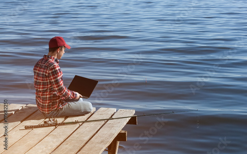 A fisherman man works on a laptop, sits on a wooden pier near the lake, next to it there is a fishing pole.