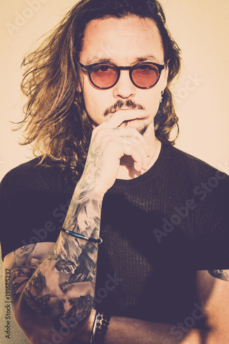 beautiful man with long black hair look at you thinking about it. Fashion model concept with vintage filter and warm tones. Black t-shirts and eyeglasses, trendy look and tattoes photo