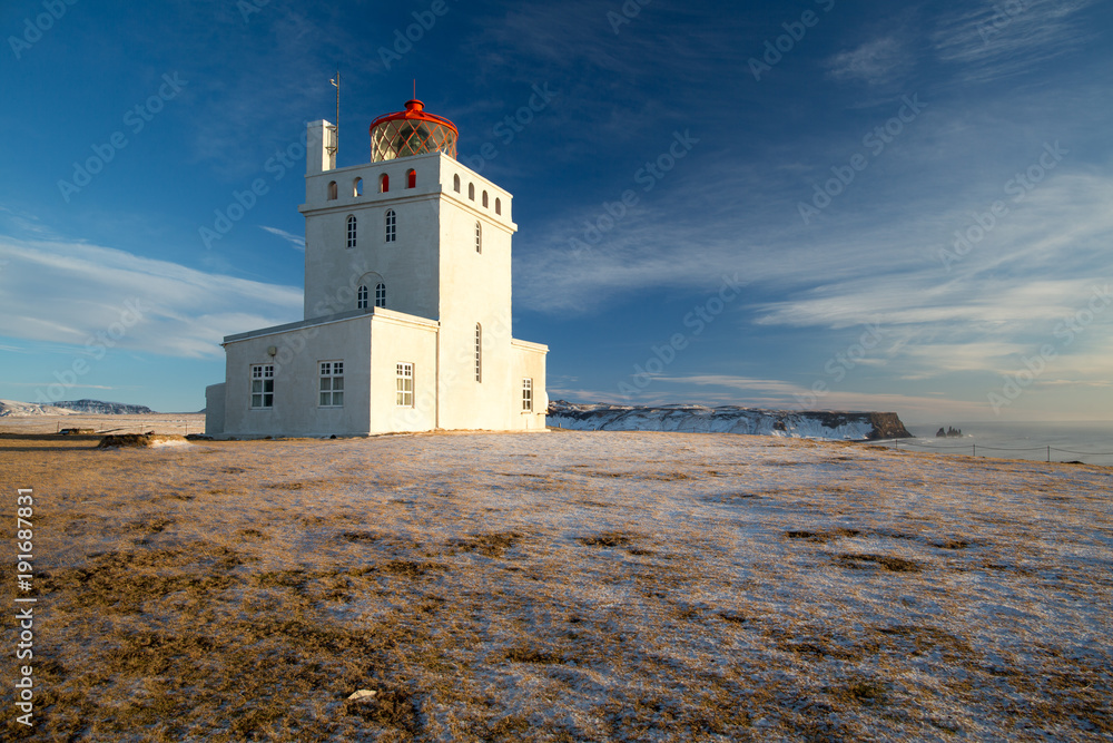 Dyrhólaey lighthouse in winter with icing on a grass. Dyrhólaeyjarviti light house located on central south coast of Iceland with famous view on Reynisfjara Black Sand Beach. Winter sunny weather.
