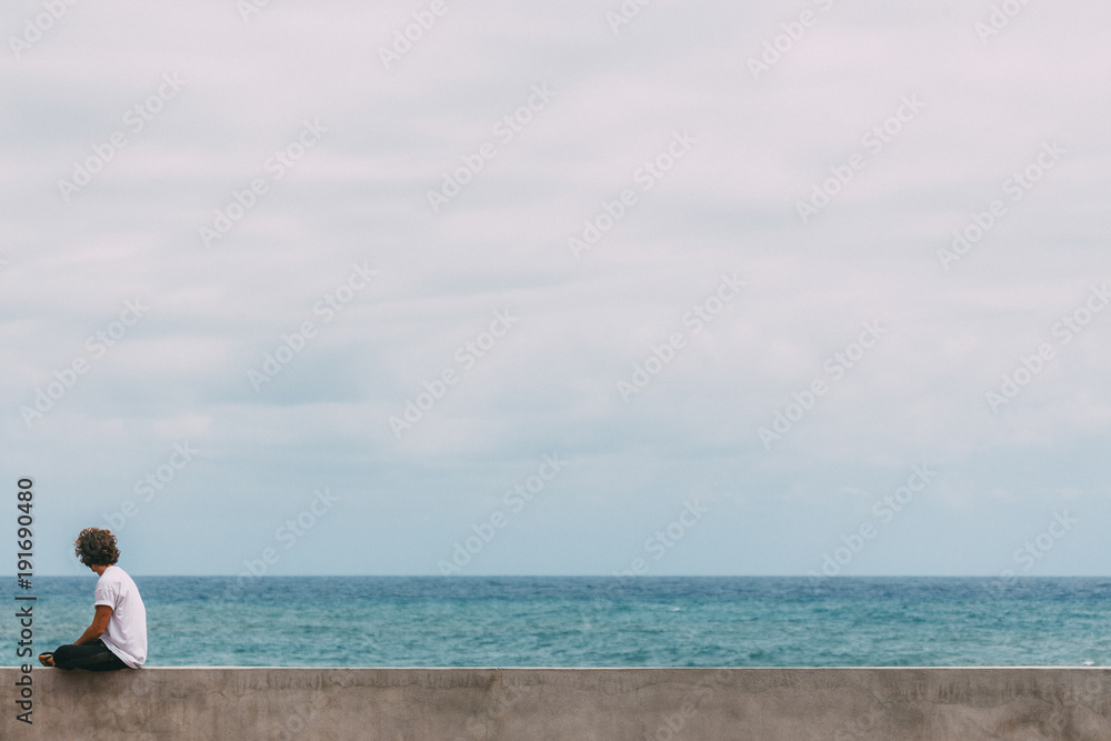 Young surfer sitting on a concrete wall with long brown curly hair in white t-shirt is watching the blue ocean and searching for waves