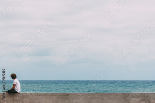Young surfer sitting on a concrete wall with long brown curly hair in white t-shirt is watching the blue ocean and searching for waves © Filip