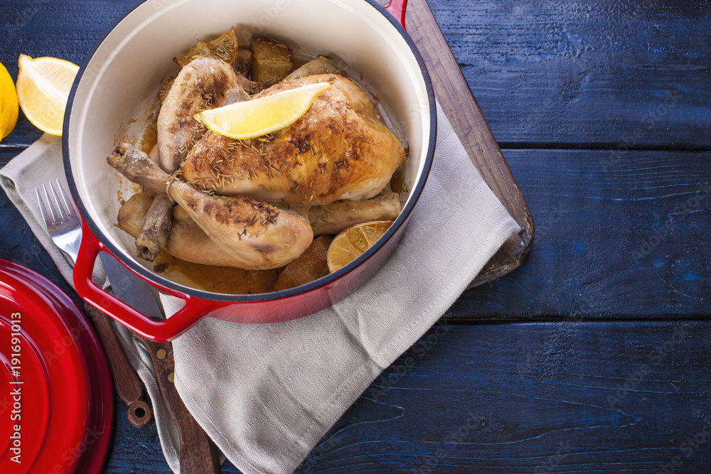The whole chicken baked with lemon and rosemary in a red cast iron. Blue wooden background and gray towel. Knife and fork. Free space for text.
