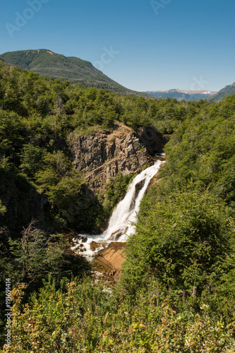 Portrait of a natural waterfall surrounded by a forest of green trees and blue sky