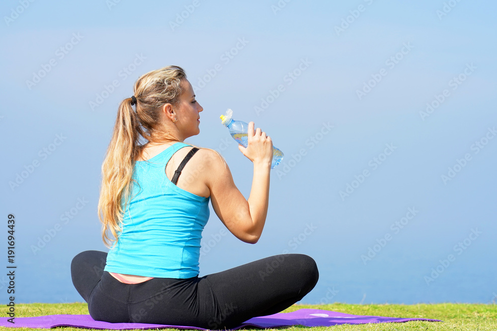 Young woman sitting after a yoga class is drinking water on a break sitting on yoga mats on the grass against the backdrop of the morning sea. Concept of health. Young woman sitting after a yoga class is drinking water on a break sitting on yoga mats on the grass against the backdrop of the morning sea. Concept of health.