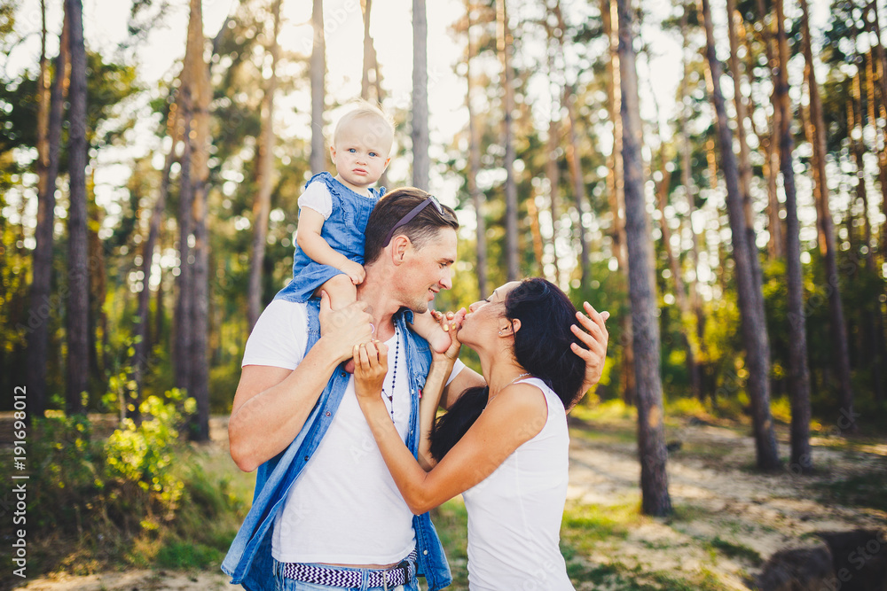 Fototapeta premium Family mom, dad and daughter sits at the daddy on the shoulders, and parents kiss on the nature in the forest in the summer at sunset