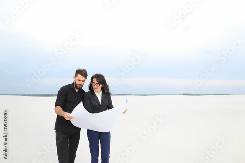 CEO male and CFO female holding whatman paper, colleagues discuss project of wind power plant in deserted place. Young woman and bearded guy standing among sands looking around. Concept of explor photo