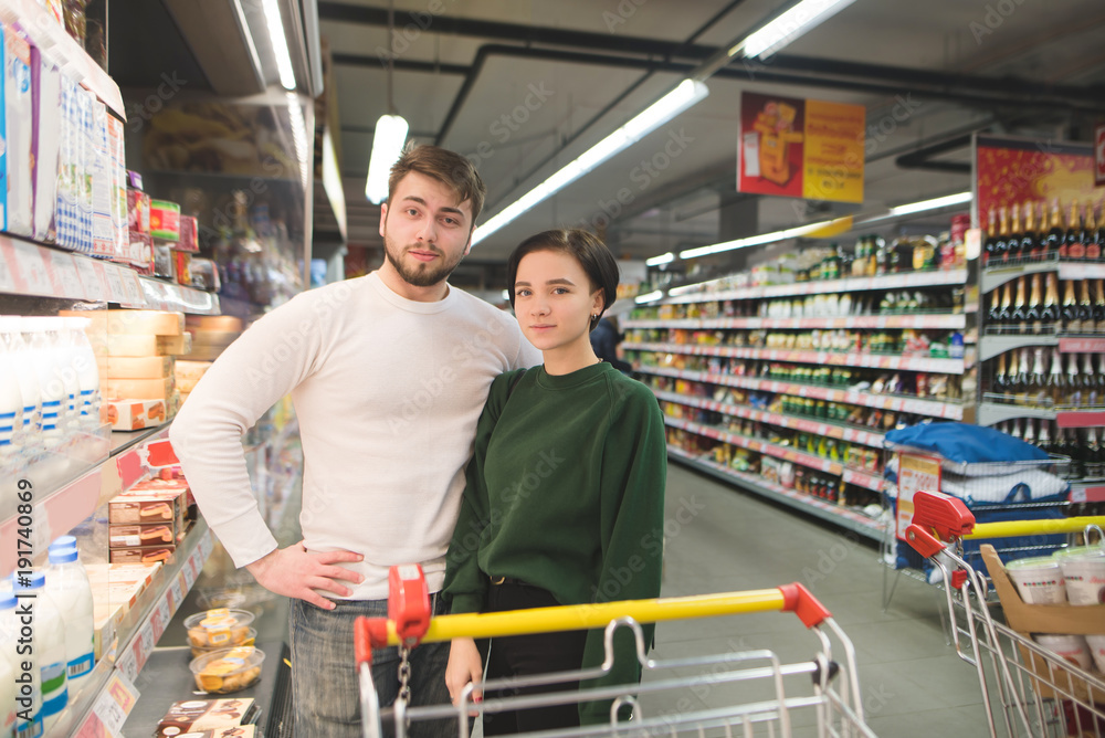 Young beautiful couple with shopping carts standing in the supermarket and looking at the camera. Family shopping at a supermarket.